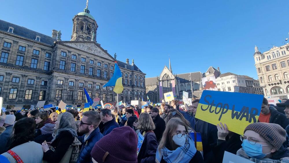 Een menigte staat op de Dam. Een van de protesteerders draagt een bord met de blauw-gele kleuren van de vlag van Oekraïne met daarop de tekst "stop war".