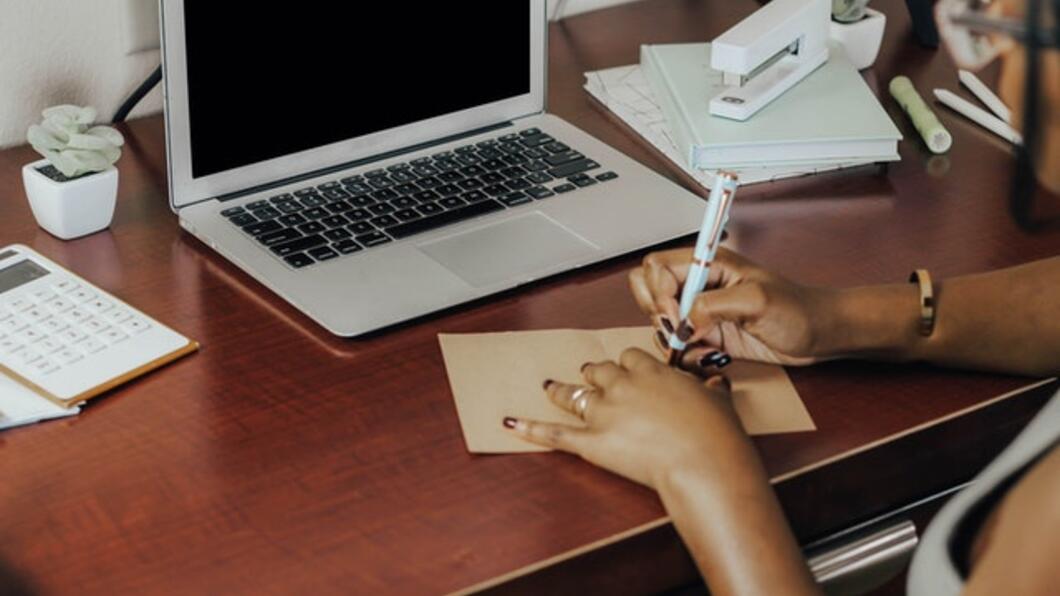 Een computer op een bureau. Voor de computer ligt een noteblok. Een bruine, vrouwelijke linkerhand ligt op het blok. Haar rechterhand houdt een pen vast.