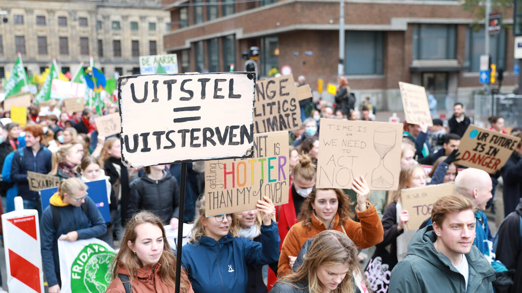 Mensen lopen in de klimaatmars. In het midden houdt iemand een protestbord vast met de tekst Uitstellen is uitsterven. 