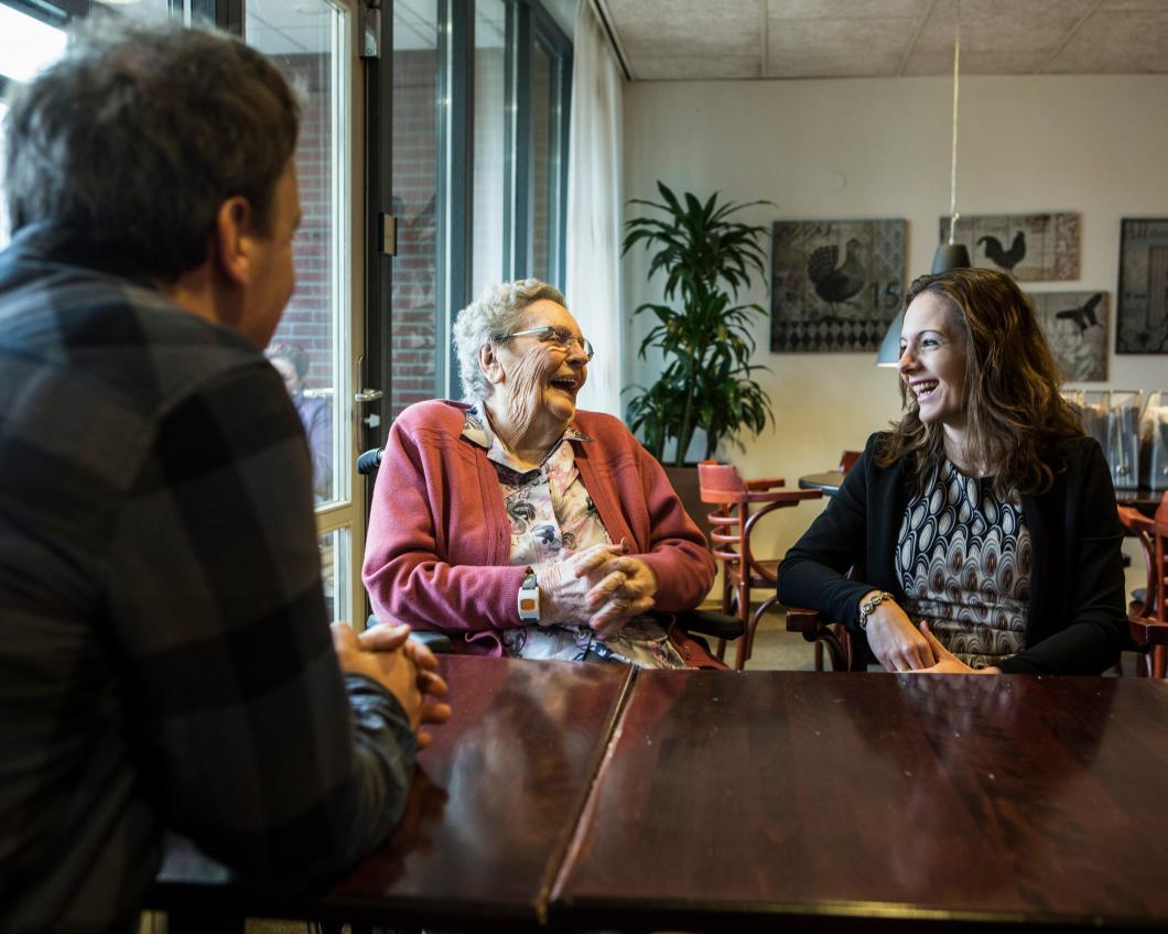 Een lachende oudere vrouw zit aan tafel met een jongere vrouw en een man