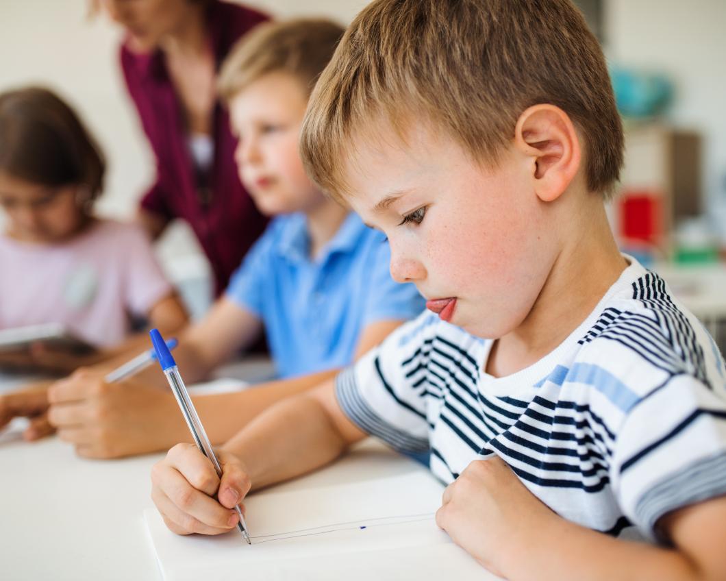 Een groep kleine kinderen in de klas die leren schrijven, met een jongetje met geconcentreerde blik vooraan in beeld