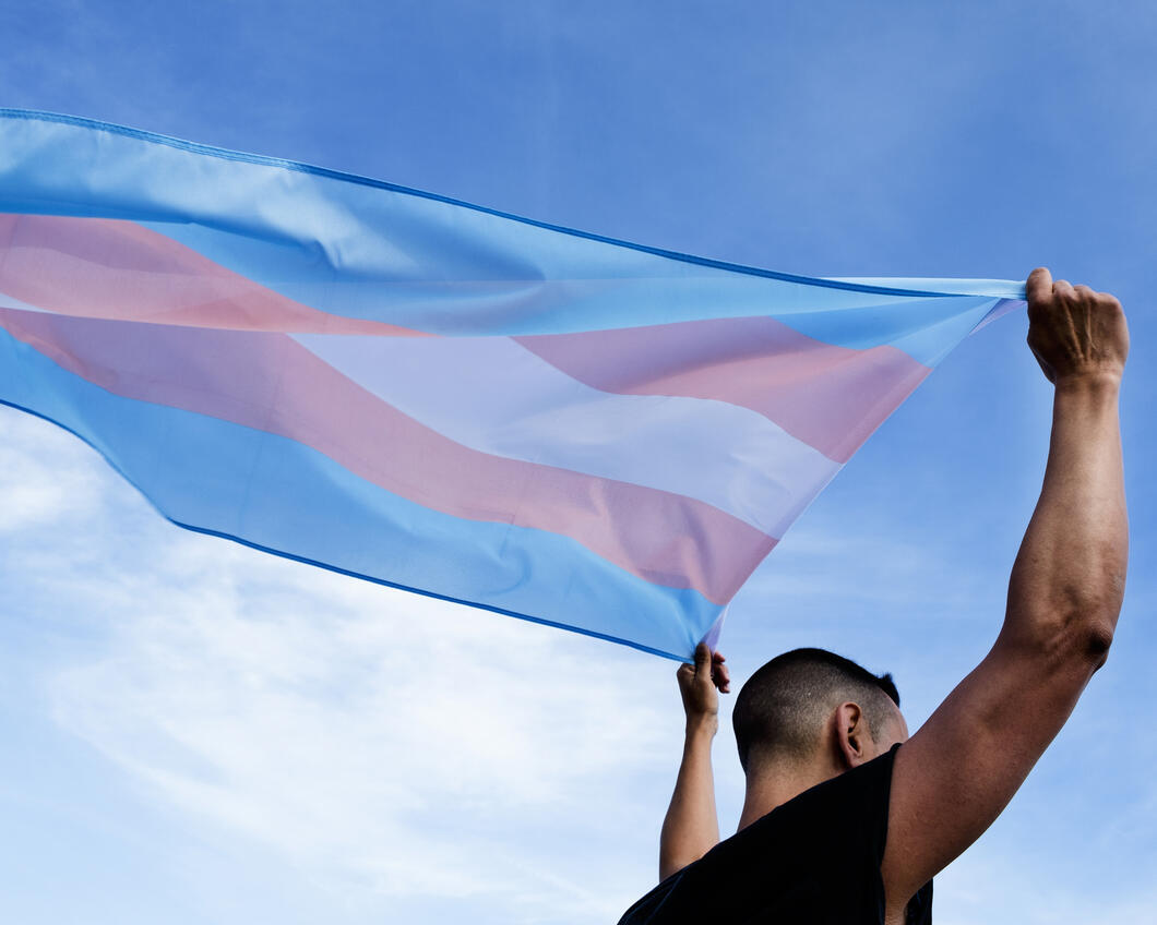 Persoon houdt de transgendervlag in de lucht. De vlag wappert in de wind.