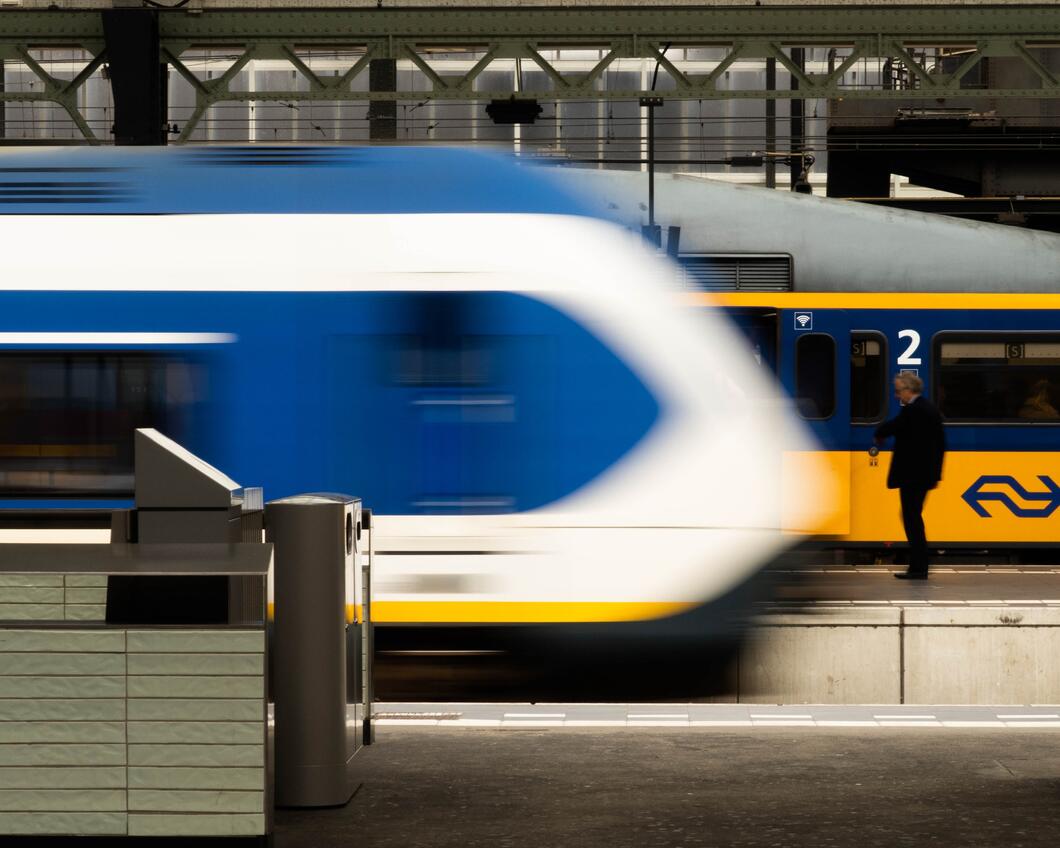 Beeld van een station, waar een sprinter voorbij rijdt terwijl een intercity trein stilstaat.