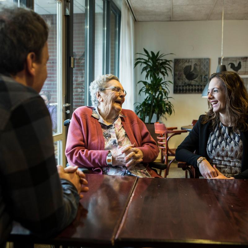 Een lachende oudere vrouw zit aan tafel met een jongere vrouw en een man