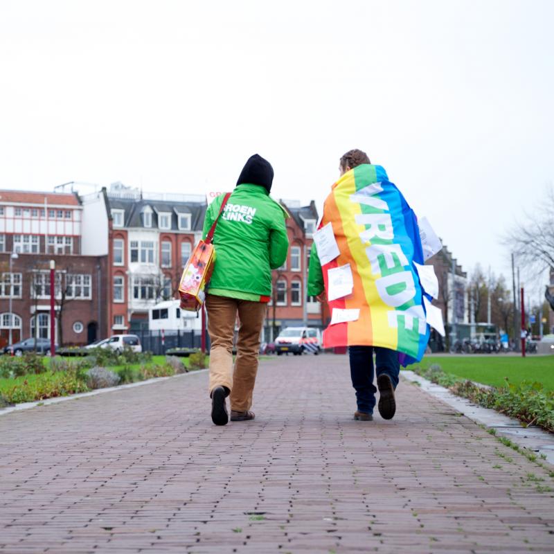 Twee vrijwilligers lopen over straat, een heeft de regenboogvlag met het woord 'vrede' over de schouders geslagen.