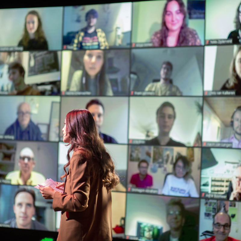 Emmelie, een vrouw met lang donker haar, staat voor een muur met een videoconferentie met allerlei mensen erop.